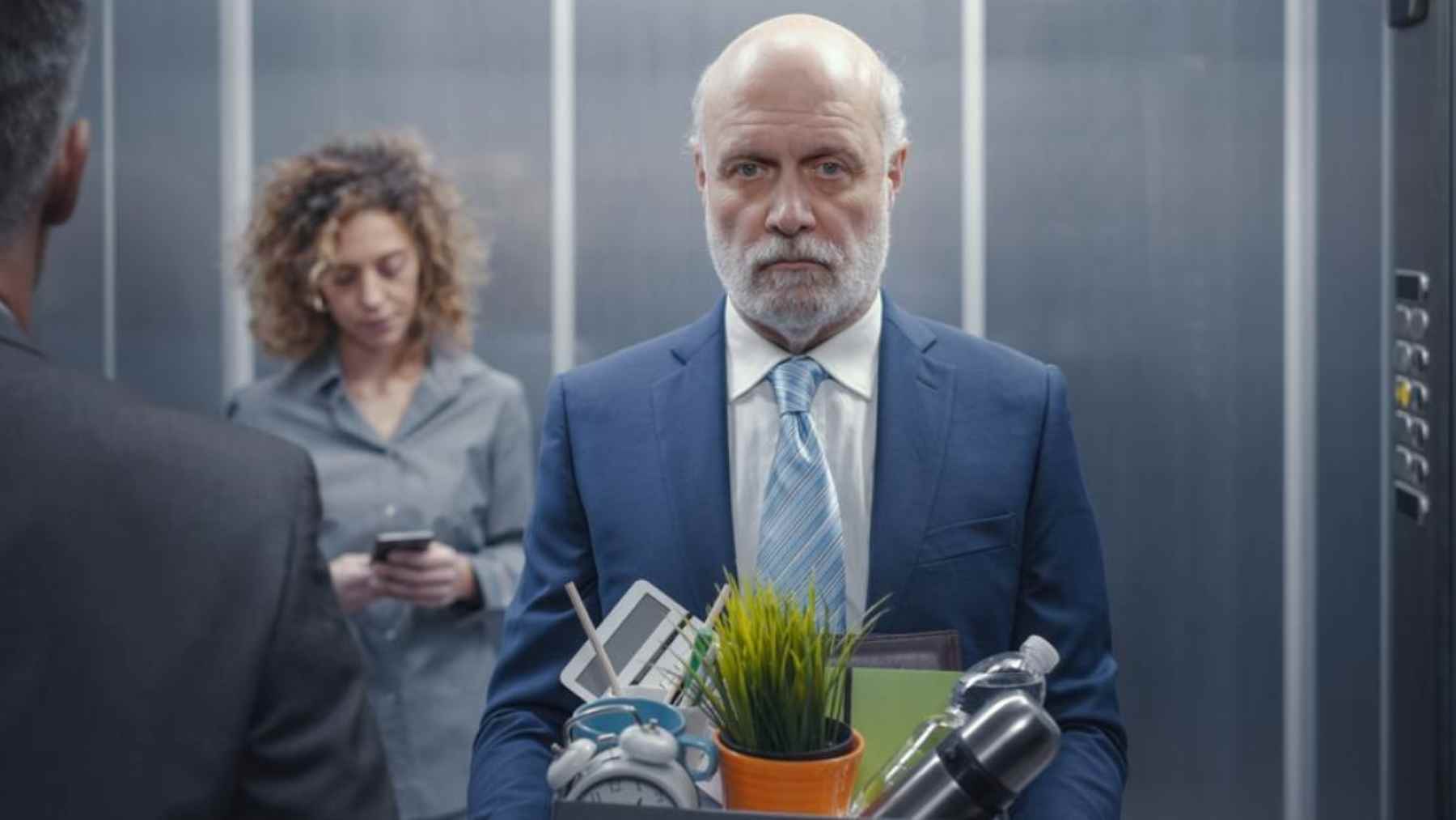An older office worker holds a box of personal items while standing in an elevator after losing his job.