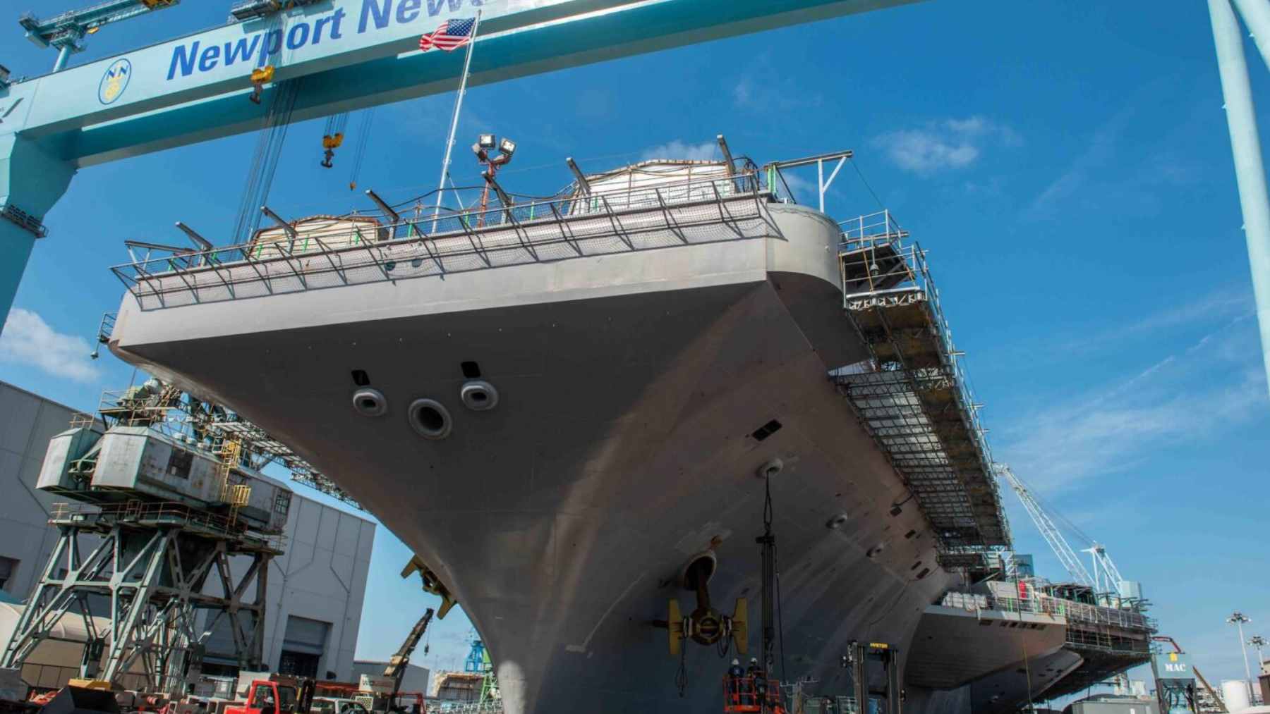 A large U.S. Navy warship under construction in a crowded shipyard with industrial cranes.