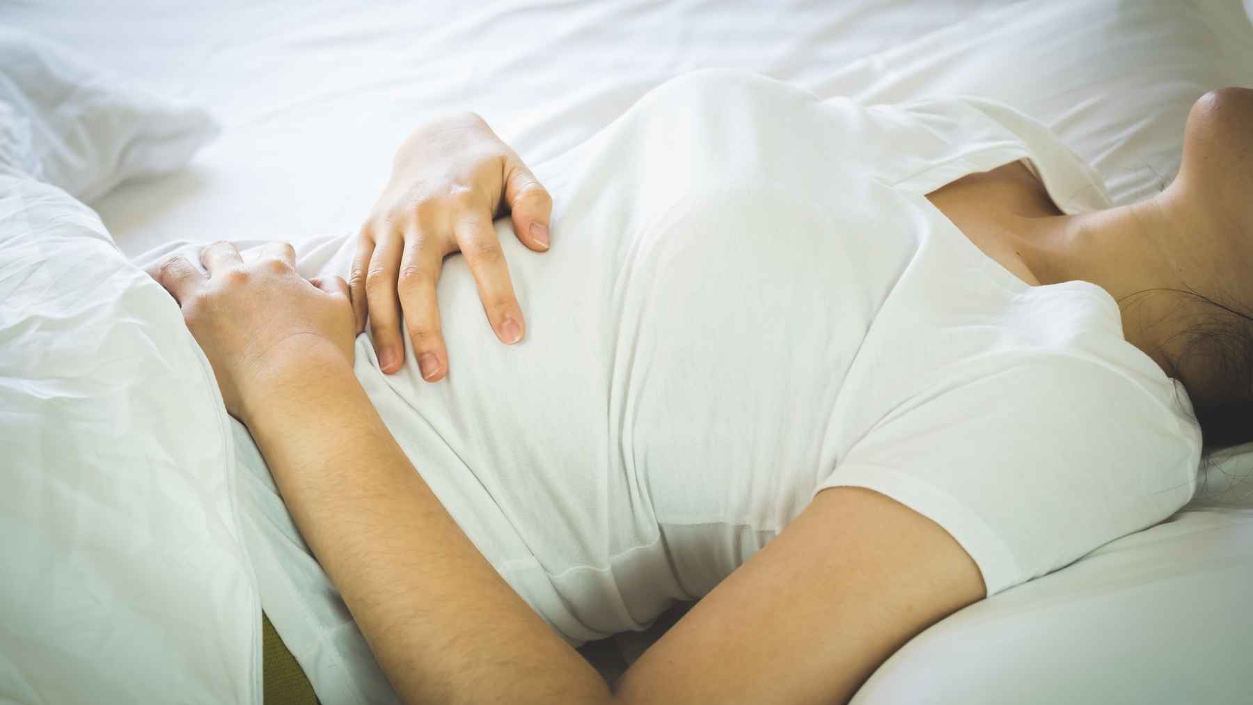 Person lying in bed holding their stomach, suggesting abdominal pain linked to digestive illness.