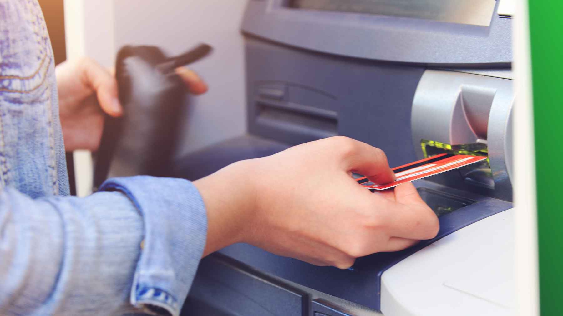 Person inserting a bank card into an ATM while holding a wallet, illustrating PIN safety and card swap theft risk.