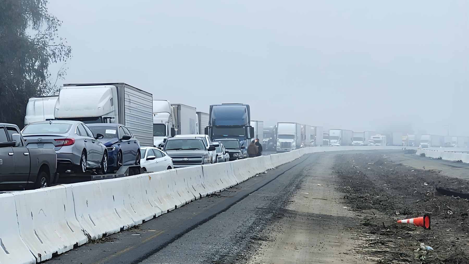A panoramic view of a massive multi-vehicle pileup on a foggy California freeway, showing crumpled cars and tractor-trailers.