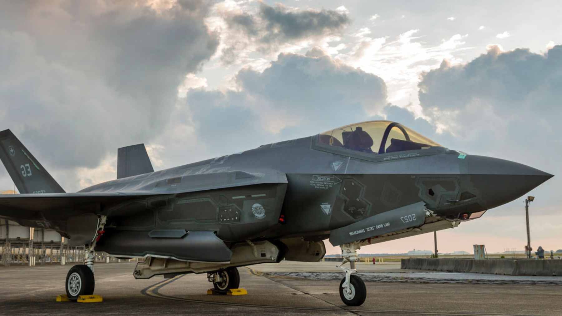 A U.S. Navy F-35C stealth fighter jet parked on a tarmac at sunset with another jet visible in the sky.