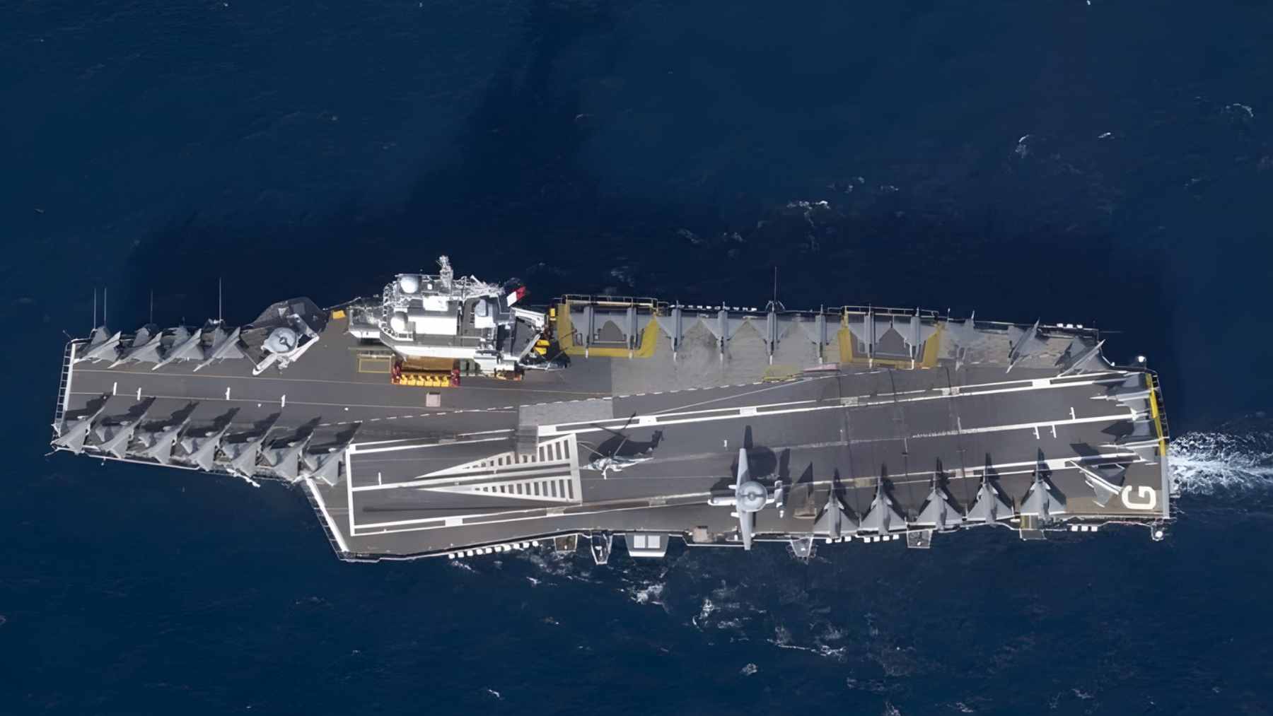 An aerial top-down view of the French nuclear-powered aircraft carrier Charles de Gaulle underway, with multiple Rafale M fighter jets parked on its flight deck.