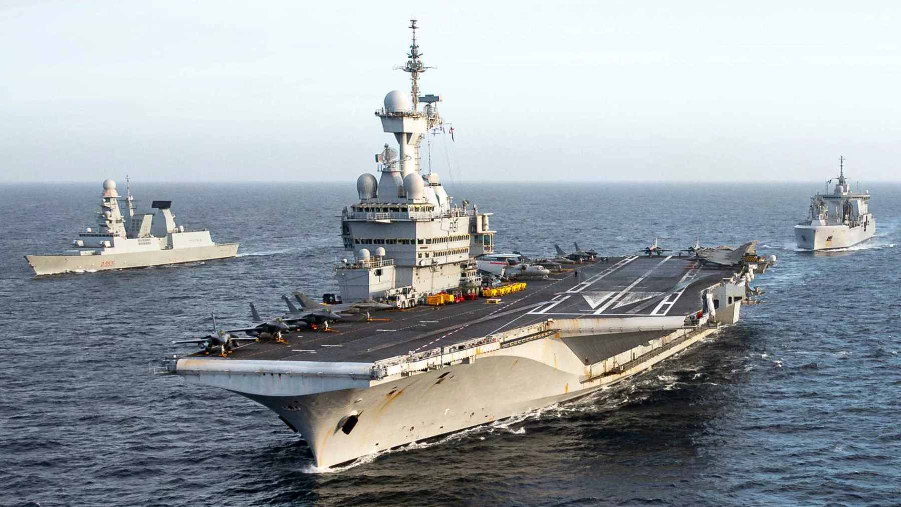 The French nuclear-powered aircraft carrier Charles de Gaulle sailing in formation with Rafale M fighter jets visible on the flight deck.