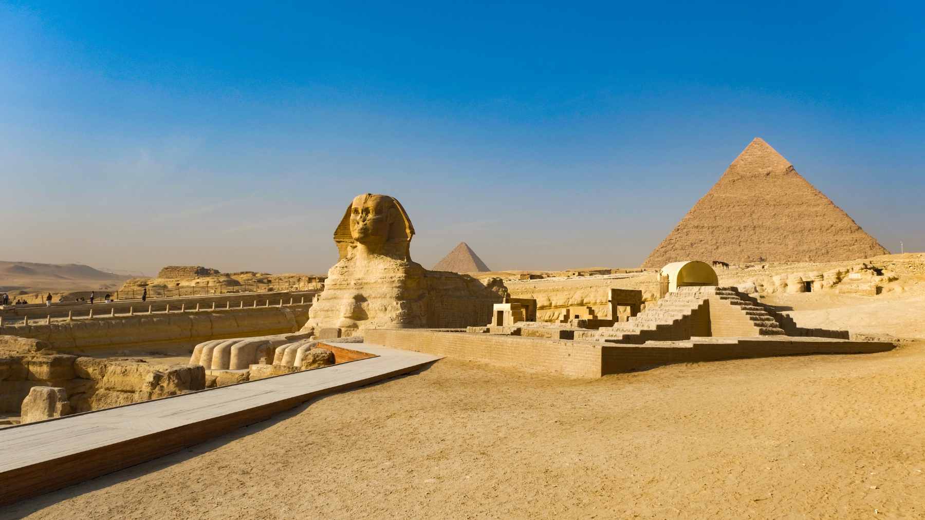 A wide view of the Giza Plateau featuring the Great Sphinx in the foreground and the Great Pyramid under a clear blue sky.
