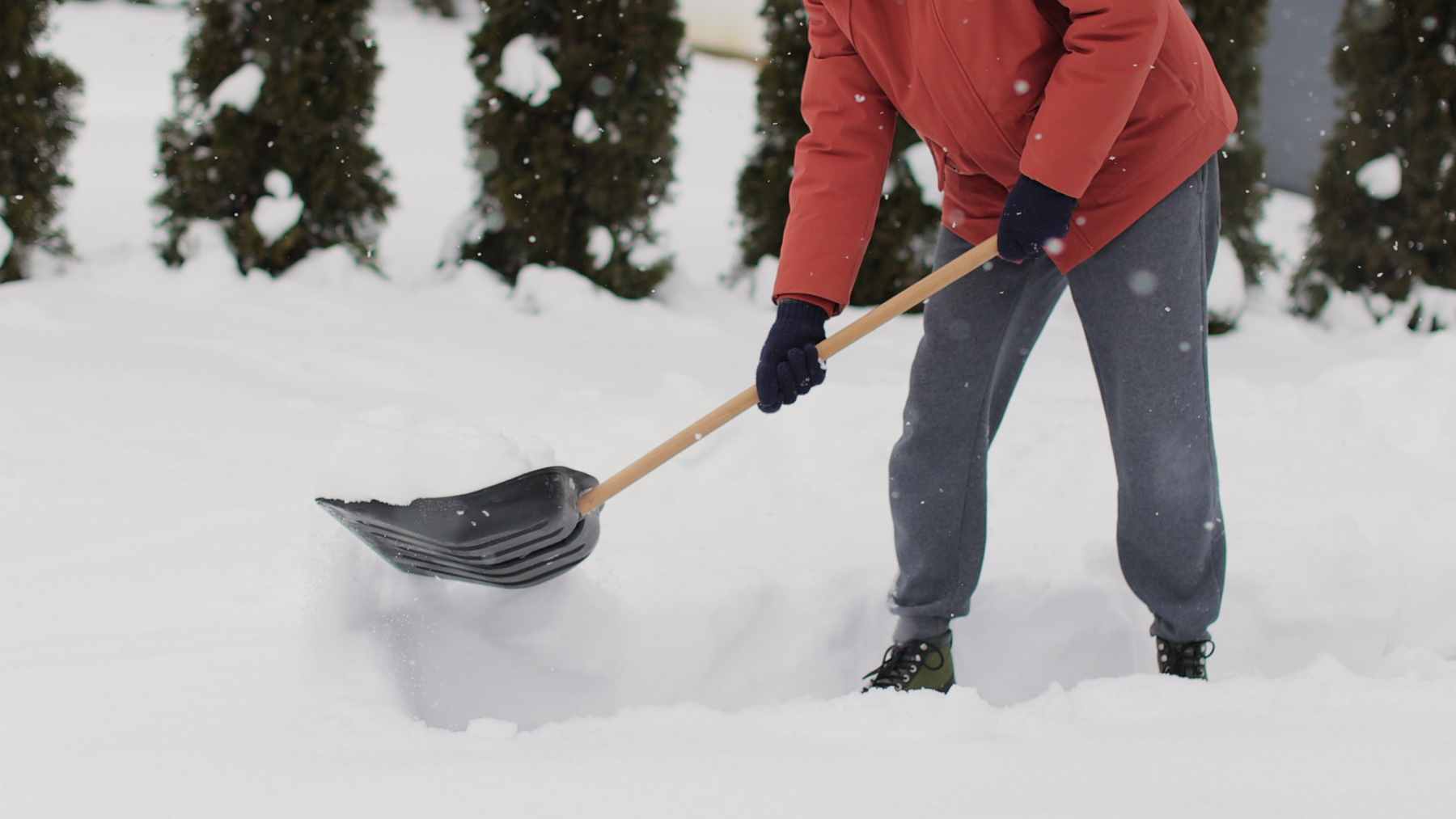 A senior man in warm winter gear using an ergonomic shovel to push light snow off a driveway.