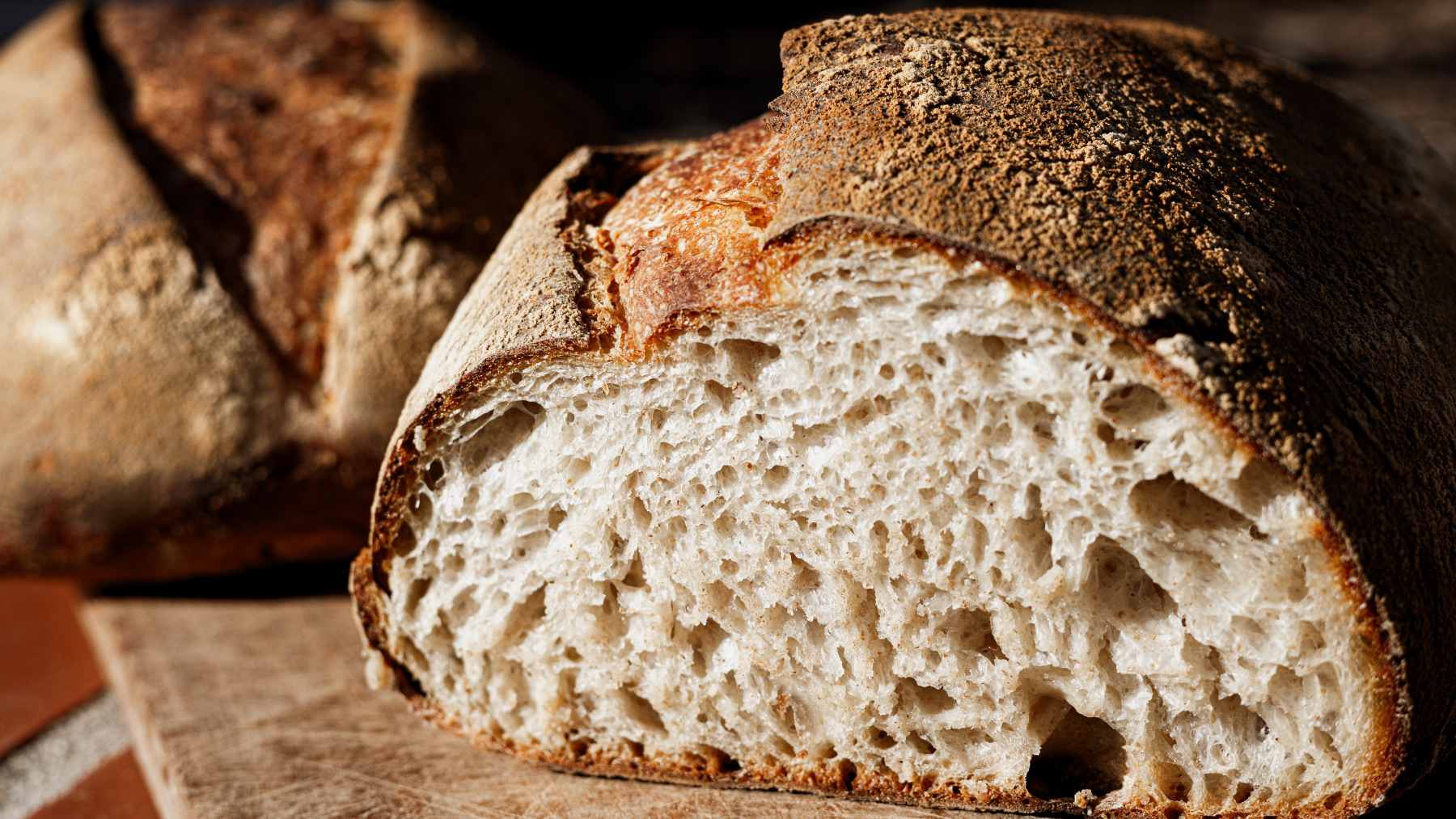 A loaf of artisanal sourdough bread sliced on a wooden board next to a glass of water and a bowl of flour.