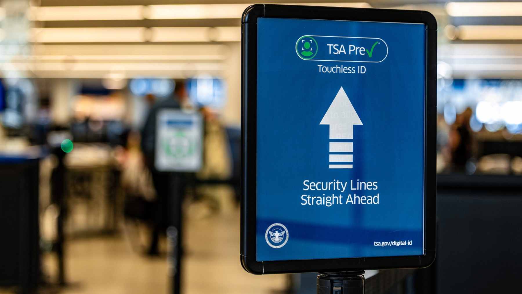 A TSA agent checking traveler documents at an airport security checkpoint next to a Real ID informational sign.