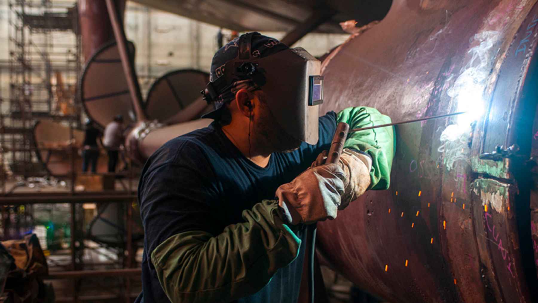 A shipyard worker welds steel on a large vessel section, illustrating the labor crunch as Navy yards compete with Amazon and Buc-ee’s.